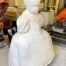 Six-axis orange industrial robot CNC-milling a life-size white foam sculpture of a girl in period dress